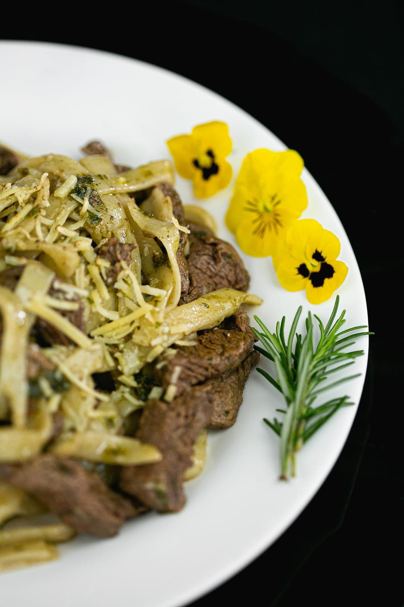 Fettuccine com tiras de carne grelhada, pesto de ervas e queijo ralado, decorado com amor-perfeito e alecrim