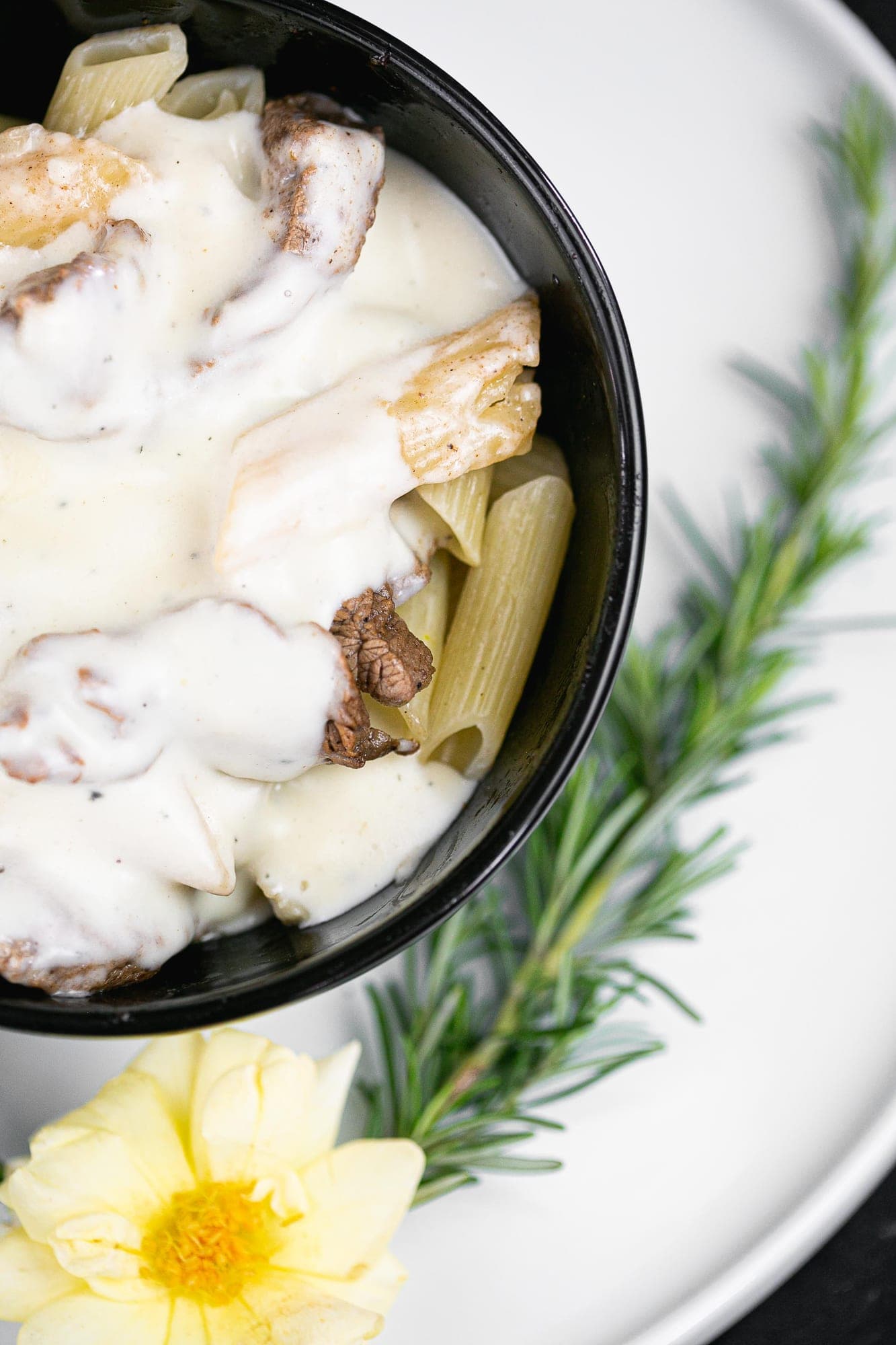 Penne ao molho branco cremoso com pedaços de carne em tigela preta, com alecrim e flor comestível